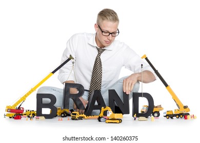 Build Up A Brand Concept: Smiling Businessman Building The Word Brand Along With Construction Machines, Isolated On White Background.