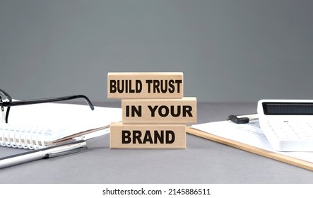 BUILD TRUST IN YOUR BRAND Text On A Wooden Block With Notebook,chart And Calculator, Grey Background