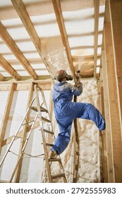 Builder applying spray foam insulation to roof and wall structure in unfinished house interior. Protective suit, ladder, professional gun, home renovation, thermal protection, modern building concept