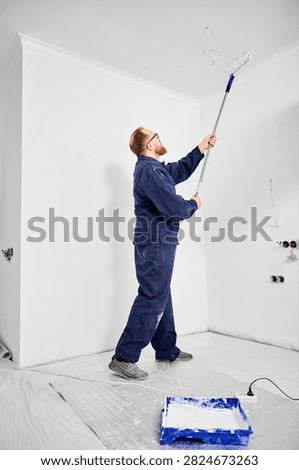 Builder applying white paint overhead using long handle roller. Clean modern interior under renovation, surface finishing and home improvement concept