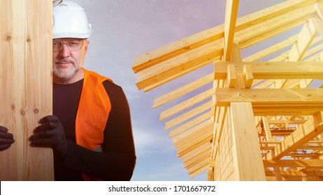 Builder With A Board In His Hands On The Background Of The Frame Of The Cottage. Construction Of A Wooden Building. Delivery Of Lumber To The Construction Site. A Carpenter In A Hard Hat.