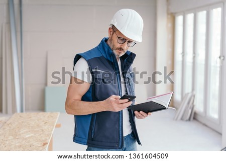 Builder checking a message on his mobile phone or making a call as he stands holding a journal on site in a new build house