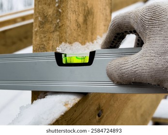 Builder checks alignment of timber with a level tool amidst snow, emphasizing precision in cold weather construction efforts
