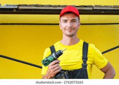 Builder with cordless drill and toolkit, standing at house under construction, at-home repairs, professional contractor business, DIY improvement skills.