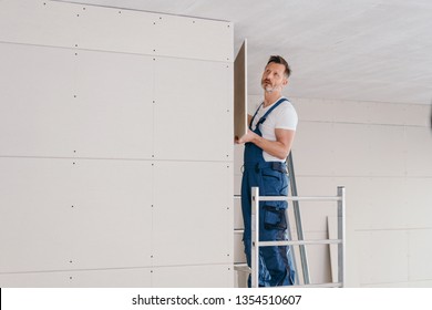 Builder In Denim Overalls Standing On A Metal Platform Fitting Wall Cladding In A New Build House Holding A Sheet In His Hands