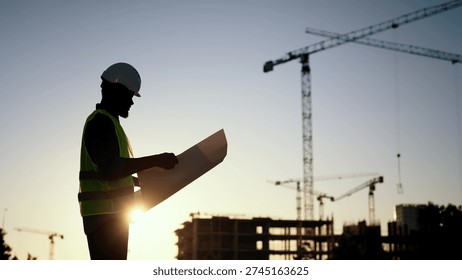 Builder, engineer or architect in helmet checking drawings at construction site. Silhouette of foreman, industrial building, engineering house industry, inspector examining plan of buildings
