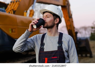 Builder In Helmet Drinking Coffee From Paper Cup On Coffee Break. Excavator On The Background.