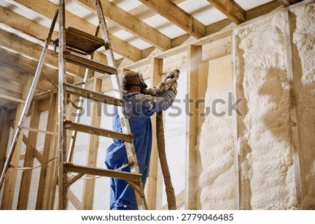 Builder insulating wall cavity with spray foam using professional plural component gun. House renovation scene with ladder and protective gear, modern construction technology and comfort concept