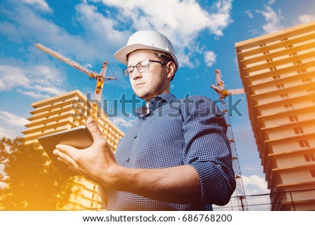 builder is man in protective helmet, glasses and tablet computer in hands amid a crane, a multi-storey building outside, on the street. Concept construction, business, technology. Sun shimmer