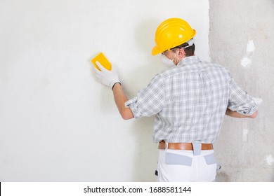 Builder Sanding And Smoothing A Newly Plastered White Interior Wall In A House Getting It Ready For Painting Wearing A Dust Mask And Hardhat