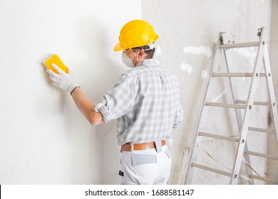 Builder sanding and smoothing a newly plastered white interior wall in a house getting it ready for painting wearing a dust mask and hardhat