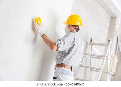 Builder Sanding And Smoothing A Newly Plastered White Interior Wall In A House Getting It Ready For Painting Wearing A Dust Mask And Hardhat