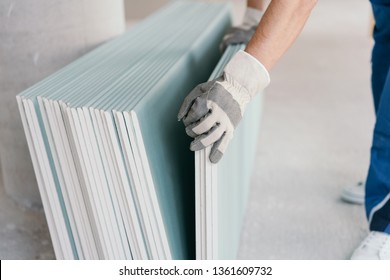 Builder Taking A Sheet Of Chip Board Cladding From A Stack Against The Wall With His Gloved Hands Indoors In A New Build Home