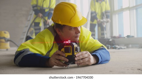 Builder in uniform lying on floor using laser level at construction site. Construction worker doing measures with laser level in renovating house