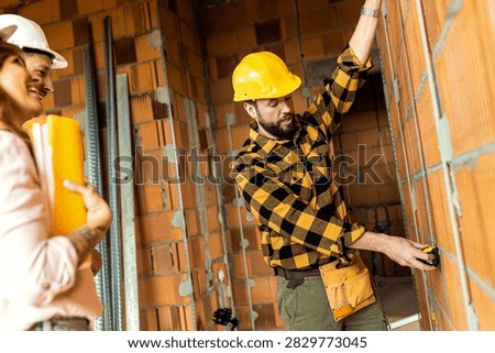Builder wearing a yellow hardhat using a tape measure against a brick wall, demonstrating precise dimensions to a smiling adult couple wearing safety helmets inside their home construction project.