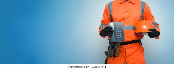 Builder Worker Is Holding In Hand A Handset Phone And Hard Hat Isolated On Blue Background With Copy Space. Call To A Repair Man Concept. Contact Us.