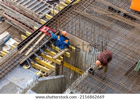 Builders in orange helmets make a steel reinforcement cage for pouring a concrete slab. Steel rods and metal formwork on a construction site. Monolithic construction. Cast-in-place concrete