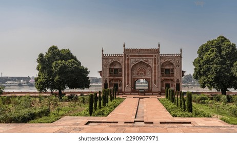 The building in the complex of the tomb of Itmad-Ud-Daula, built of red sandstone, by the river. Arches, spires, white ornaments are visible. Green vegetation on the sides of paved footpaths. India. 