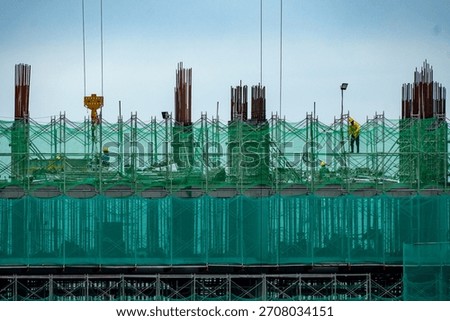 Building construction in progress, featuring workers, green safety netting, and exposed rebar against a light sky.