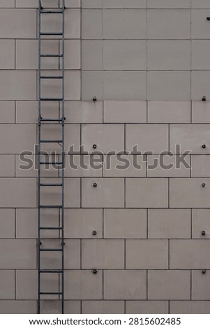Building facade featuring beige rectangular cladding panels, exposed bolts, and a dark metal fire escape ladder.