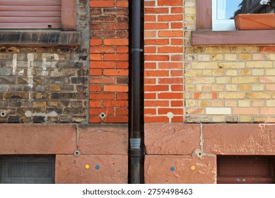 Building facade split by a vertical drainpipe: the left side shows older, darker bricks with wear and graffiti and a closed shutter; the right side features newer, lighter bricks with a mirrored window and flower box. A red sandstone base contains small colored circular markers and metal grilles. The image highlights contrasting textures, tones, and signs of renovation or adjoining properties.