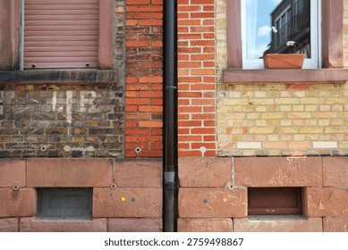 Building facade split by a vertical drainpipe: the left side shows older, darker bricks with wear and graffiti and a closed shutter; the right side features newer, lighter bricks with a mirrored window and flower box. A red sandstone base contains small colored circular markers and metal grilles. The image highlights contrasting textures, tones, and signs of renovation or adjoining properties.