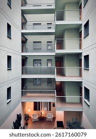 The building features multiple levels with light green walls, minimalist railings, and wooden doors, each leading to individual units. The balconies overlook a peaceful ground floor lounge area.
