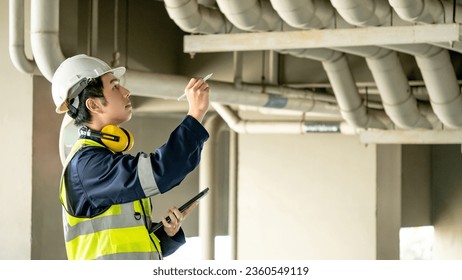 Building inspector man using digital tablet pointing at plumbing pipeline system. Asian male worker in green vest, protective ear muffs and safety helmet working for building maintenance inspection - Powered by Shutterstock