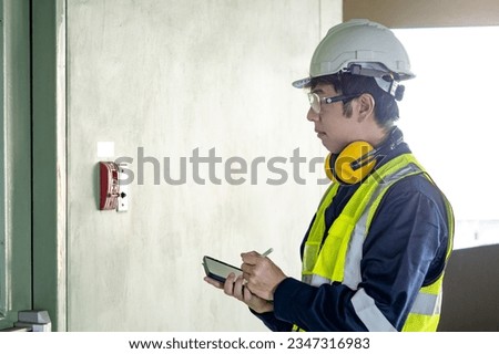 Building inspector man using digital tablet checking wall mounted fire alarm station near fire exit door. Asian male worker in reflective vest and helmet checking door hardware for safety and security