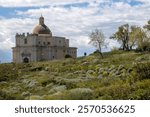 Building of the Old Cathedral (Duomo Vecchio) in the area of Milazzo Castle (Castello di Milazzo), surrounded by fresh greenery in the spring. Blue sky, white clouds. Milazzo, Sicily, Italy.