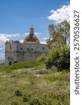 Building of the Old Cathedral (Duomo Vecchio) in the area of Milazzo Castle (Castello di Milazzo), surrounded by fresh greenery in the spring. Blue sky, white clouds. Milazzo, Sicily, Italy.