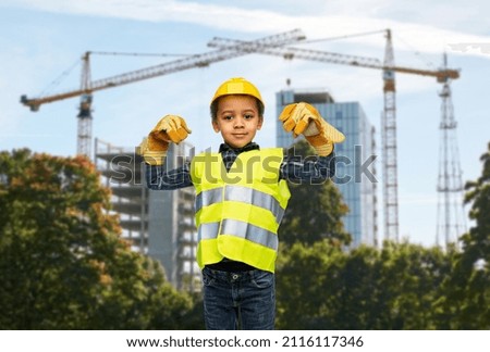 building and profession concept - smiling little boy in protective gloves, yellow safety vest and helmet showing his power over construction site background
