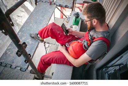Building site worker sitting high on the scaffolding, resting and using mobile phone