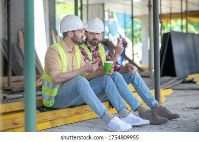 Building site. Building workers in yellow vests and helmets sitting on boards, having coffee with sandwiches, discussing something - Powered by Shutterstock