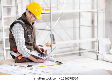 Building Supervisor In Hardhat Perched On A Workbench With Blueprints Working On His Laptop In A Building Under Construction