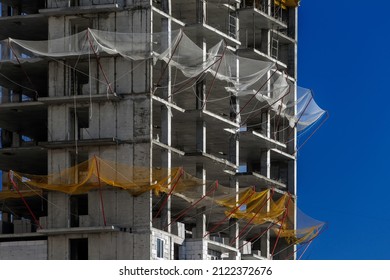 Building Under Construction Against Blue Sky. View Of The Construction Of A Modern Monolithic House With A Safety Net For Fall Prevention Fence. Housing Construction, The Growth Of New Block Houses.