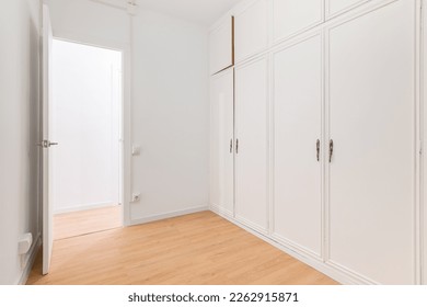 Built-in spacious wardrobes with white paneling and door open to another room with wooden laminate flooring. Concept of organizing storage and laconic interior