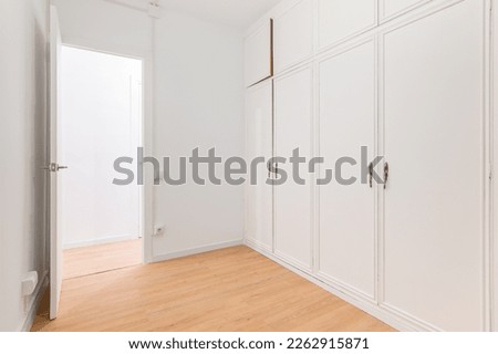 Built-in spacious wardrobes with white paneling and door open to another room with wooden laminate flooring. Concept of organizing storage and laconic interior