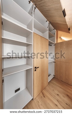 Built-in storage system featuring open wood-paneled cabinets and clean white shelves in a minimalist interior. Hardwood floors and concealed hinges. No logos visible