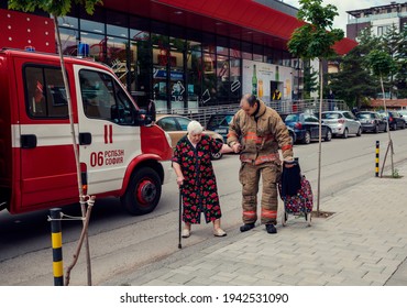 Bulgaria, Sofia, MAY 29, 2020: Fireman Helping Old Woman To Cross The Street During The Incident With Burning Car  Translation On A Vehicle - 6th Fire Brigade Sofia, On A Shop Window - Insuarance