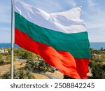 The Bulgarian flag waves proudly above a coastal city, with the vast sea and green landscape in the background. A symbol of national pride and beautiful natural scenery