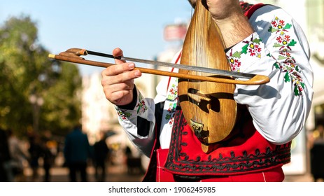 Bulgarian folk musician - violinist in traditional national costume plays an old stringed instrument - gadulka Blurred city street in background. Plovdiv Bulgaria. Bulgarian folklore and culture