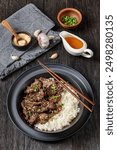 Bulgogi, Korean BBQ Beef, fried thinly sliced beef sprinkled with sesame seeds and spring onion served with long grain rice in black bowl on dark oak wooden table, vertical view