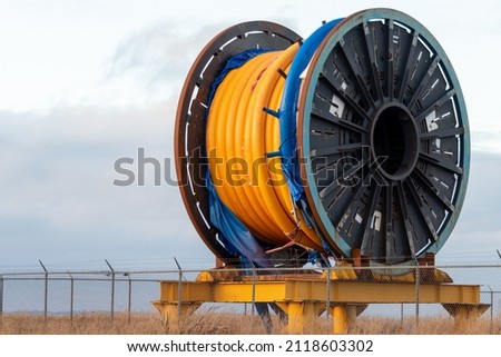 Bulk sub-sea industrial glass fiber optic cable on a metal spool on a ship's stand. The orange data line is coiled around a black reel in a storage yard. Internet communications spool storage yard.