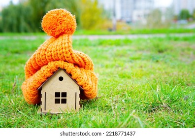 Bulky Knitted Hat Put On Top Of A Toy Wooden House Model Placed On A Green Lawn Outdoors. Staying Warm Without Cranking The Heater.