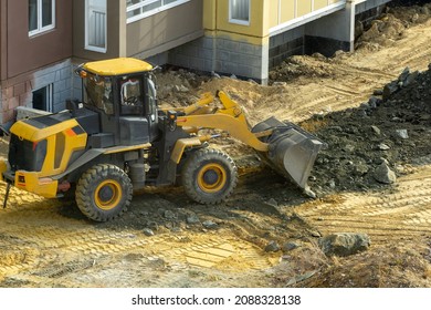 A Bulldozer Is Leveling Gravel On A Construction Site Of A Residential Building. A Yellow Tractor With A Bucket Drives On A Sandy Area Leaving Traces Of The Tread.