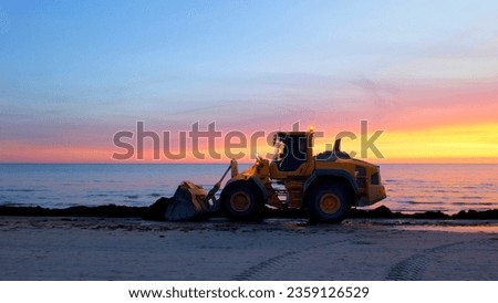 Bulldozer on the beach at sunset. A large tractor cleans the beach of sea grass. Bulldozer in motion and blur.