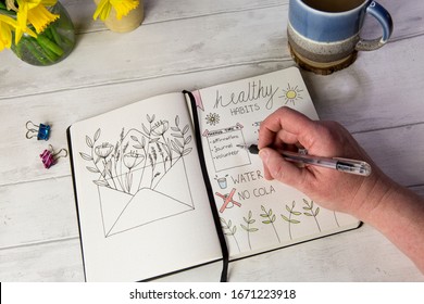 Bullet journal open on self care healthy habits layout pages with hand holding pen. Over shoulder view, fresh white table background, flowers, positive mental health message.