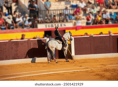 A bullfighter on horseback holds a lance in a bullring, facing the arena wall with the audience in the background, during a bullfighting event. Berja,Spain-August 3, 2024; - Powered by Shutterstock