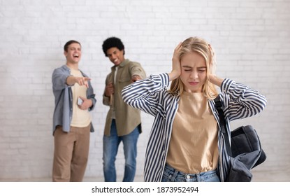 Bullying And Peer Relationships. Focus On Blonde Teen Lady With Backpack Covering Her Ears, Not To Hear Taunts From Multiracial Male Students In School Or College, On White Brick Wall Background